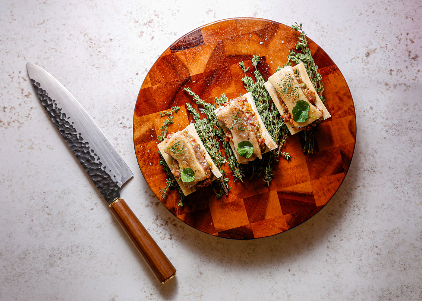 Katto Chefs Knife resting alongside a chopping board holding steak tartare served in bone marrow troughs, topped with bone marrow and thyme.